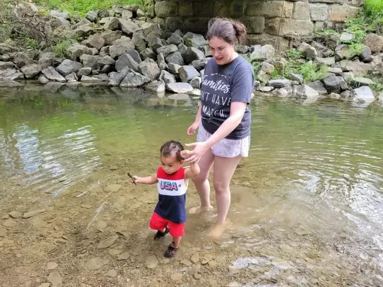Woman and child wading in shallow water of a stream; woman holds child's hand.