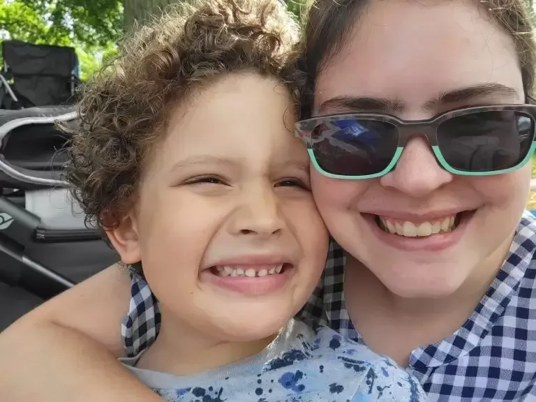Smiling woman and child outdoors, hugging close. Child has curly hair. Both are happy.