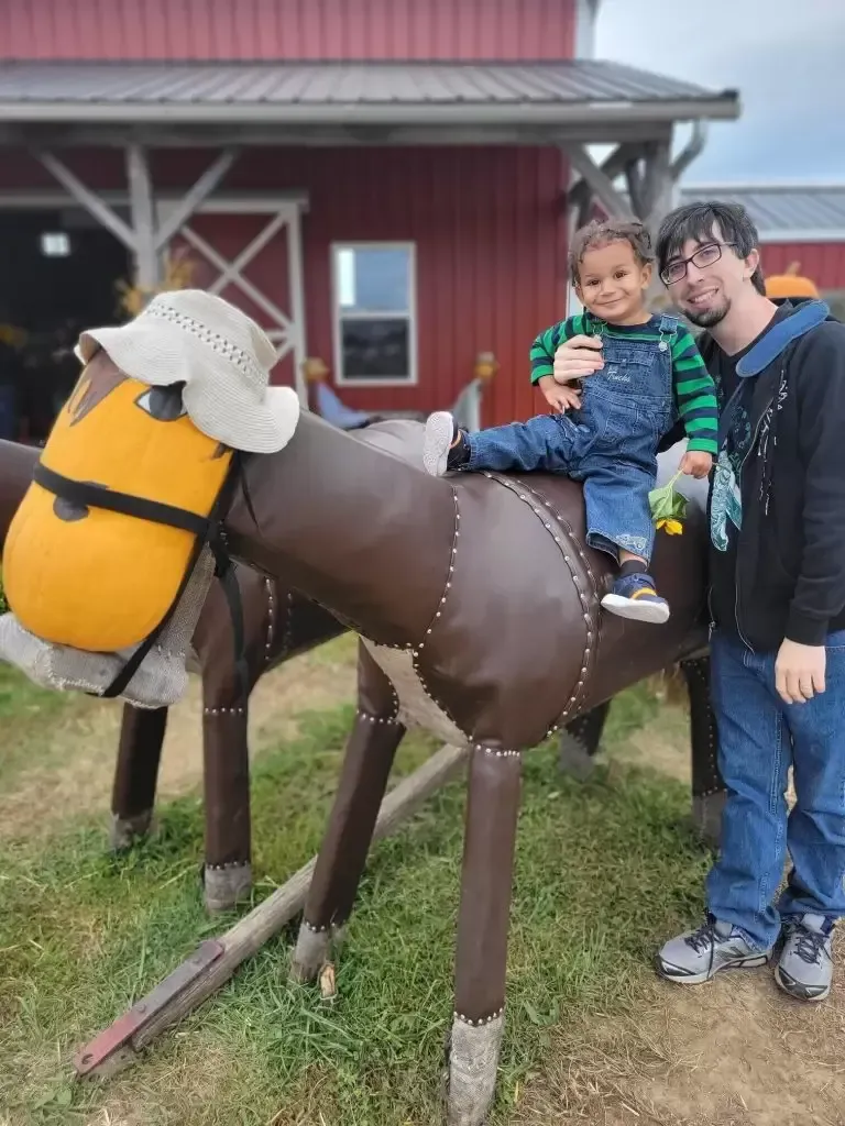 A man and child pose with a brown metal horse sculpture at a farm, the child seated on the horse.
