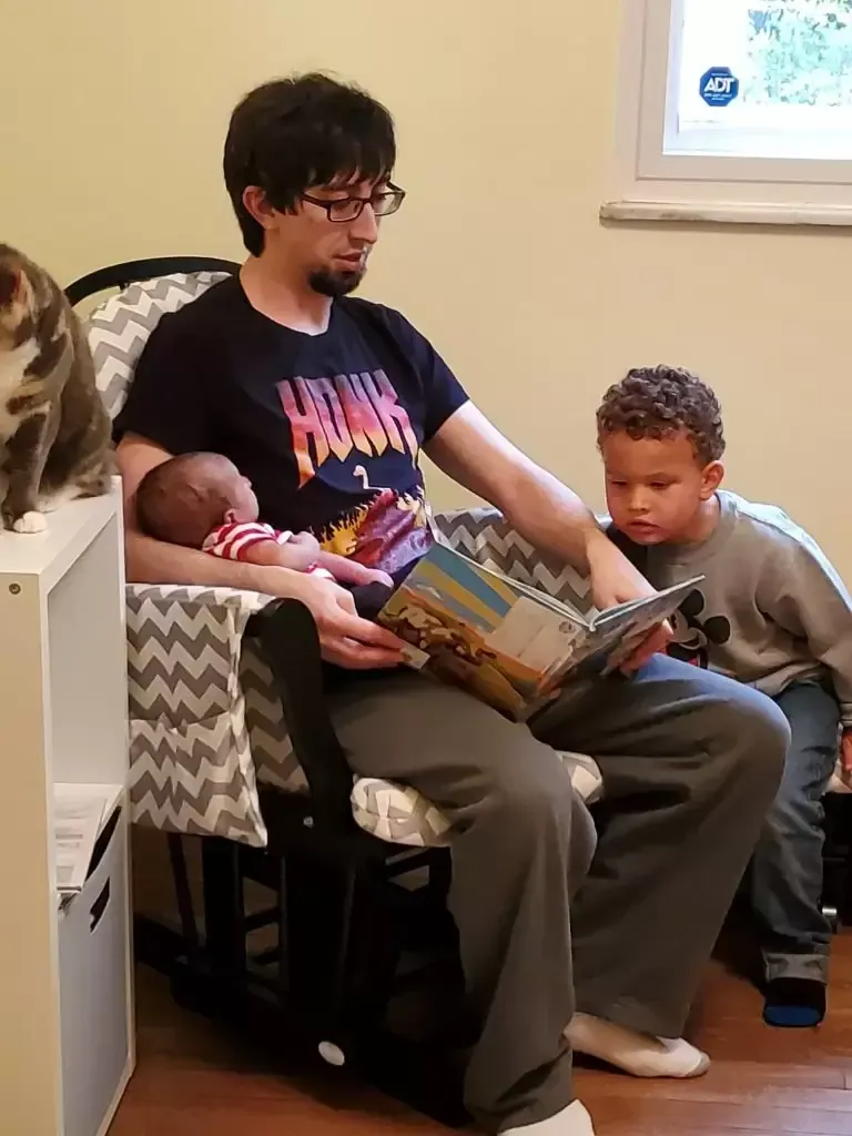 Man reads to two children, baby in arms, in a rocking chair indoors.