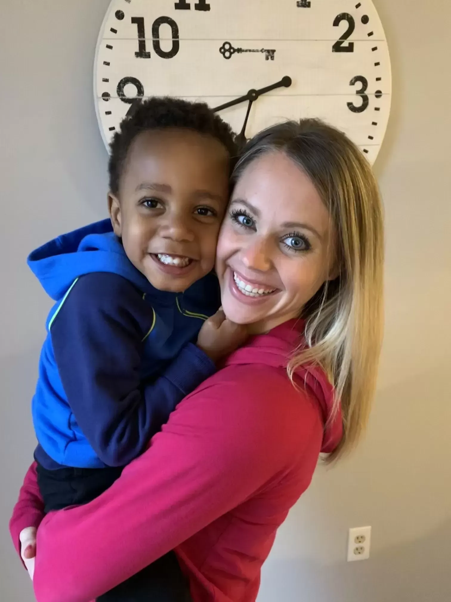 Woman holding a smiling child; both are facing the camera near a clock.