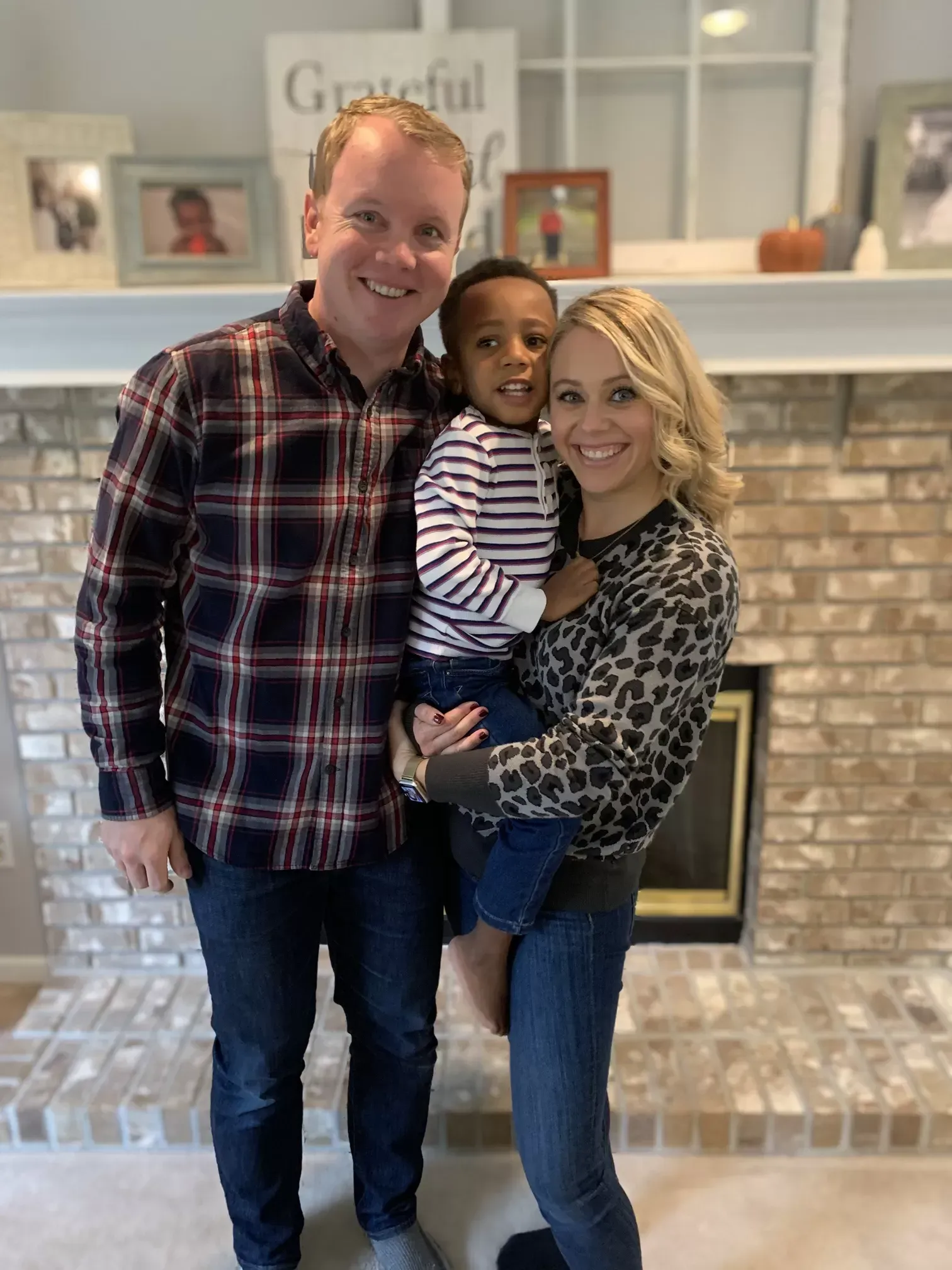 Family of three smiling in front of a fireplace. Man in plaid, woman in leopard print, holding a child.