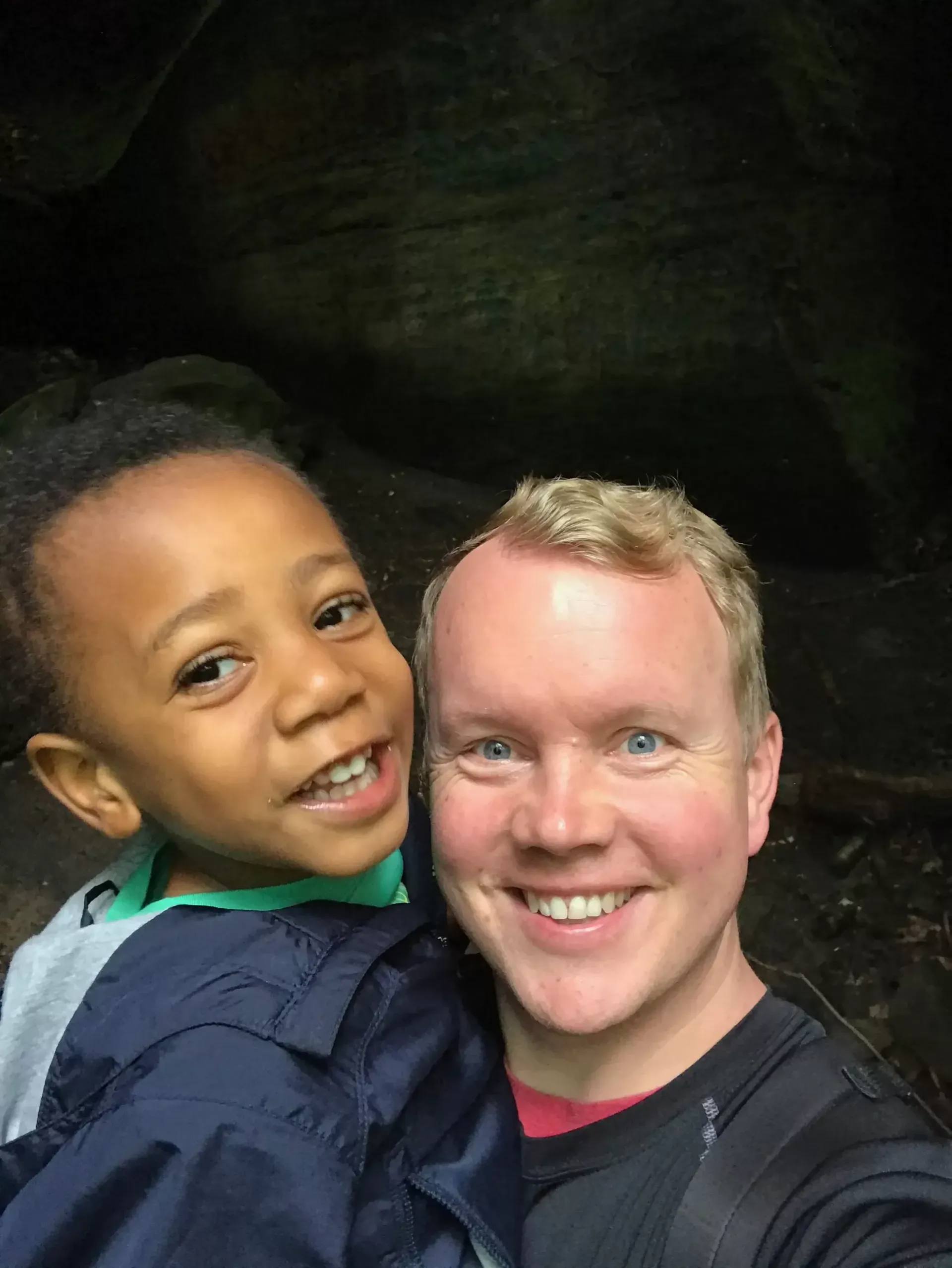 Man with fair skin smiles with a Black child, both posing close with a dark background.