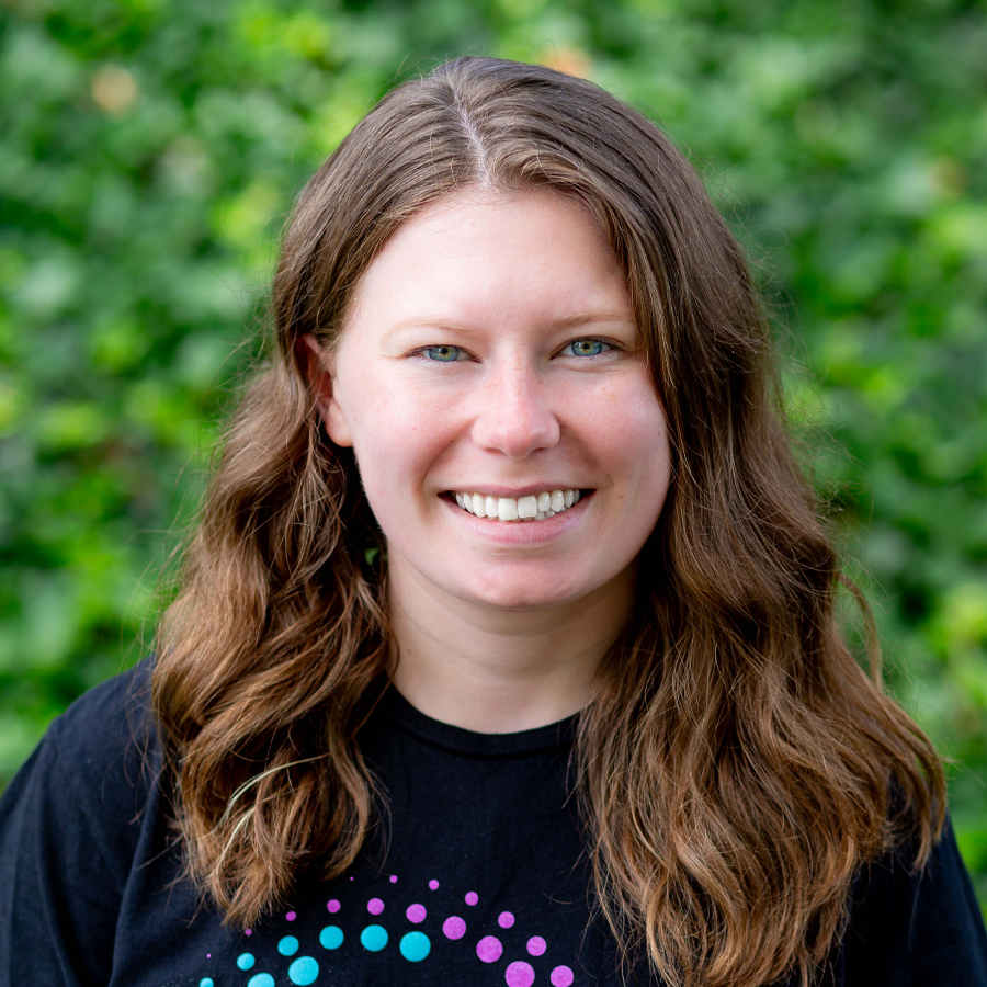 Woman with long brown hair smiles, wearing a black shirt, in front of green foliage.