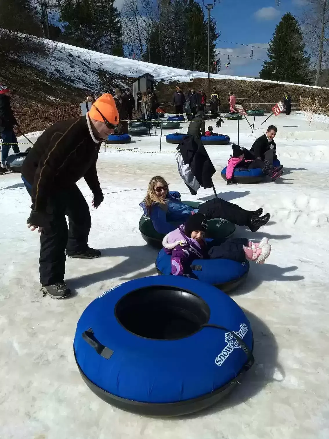 People snow tubing down a snowy hill. Man assisting, others ready to slide. Blue tubes prominent.