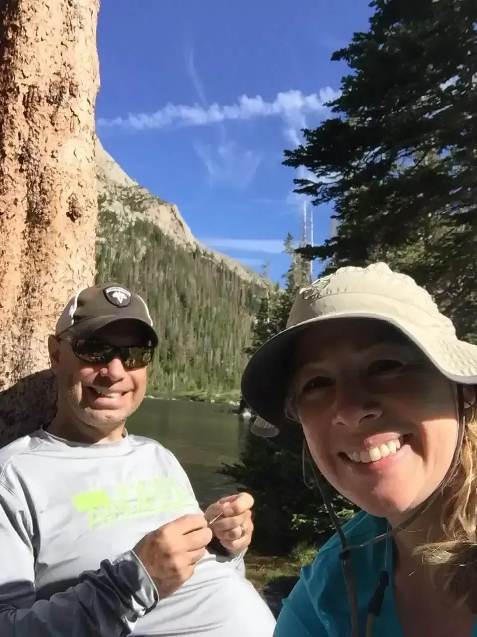 Couple smiles for a selfie by a mountain lake; man wears sunglasses, woman wears a hat; sunny day.