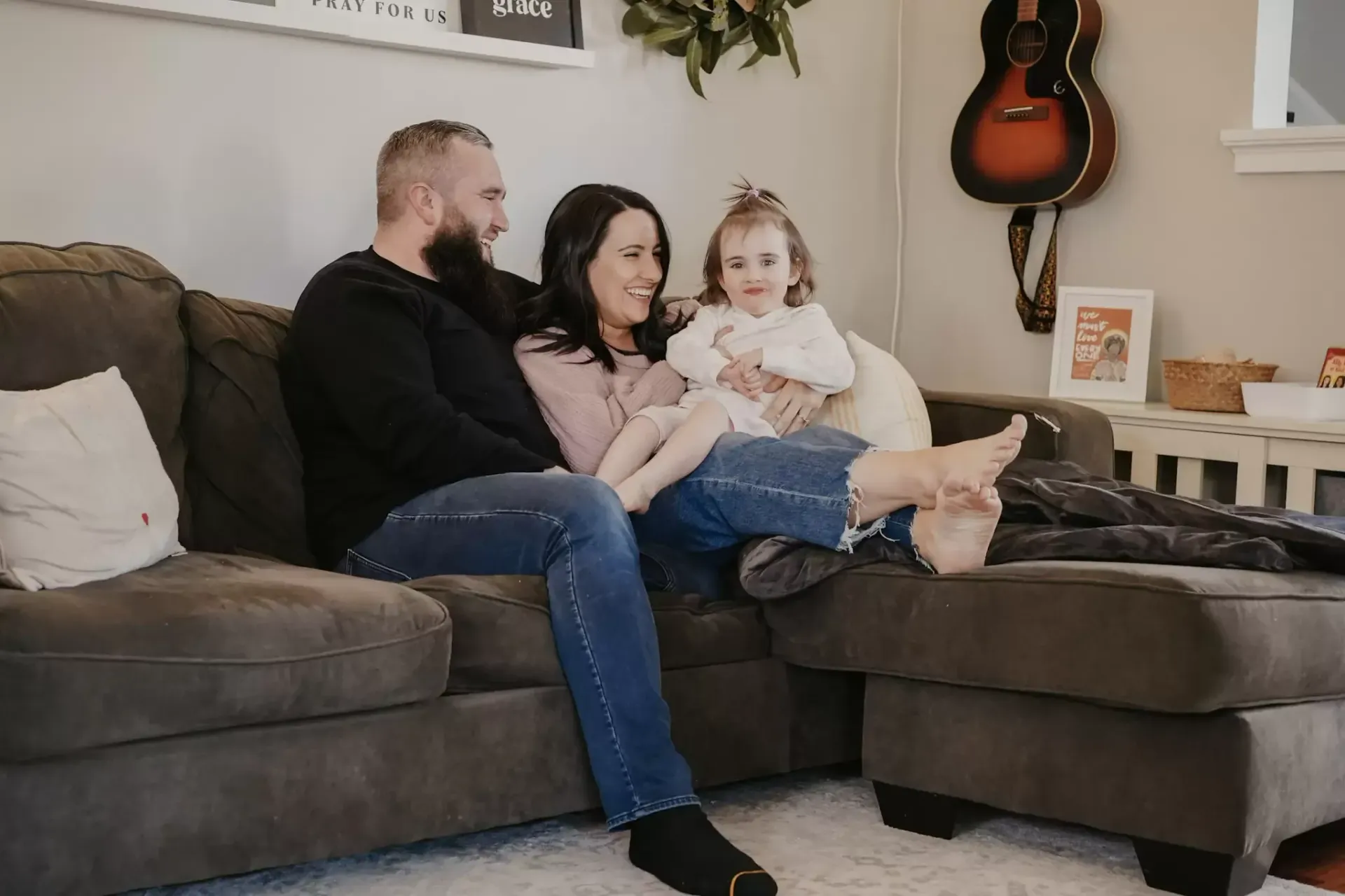 Family of three sits on a couch in a living room, smiling. A father, mother, and small child are relaxing.