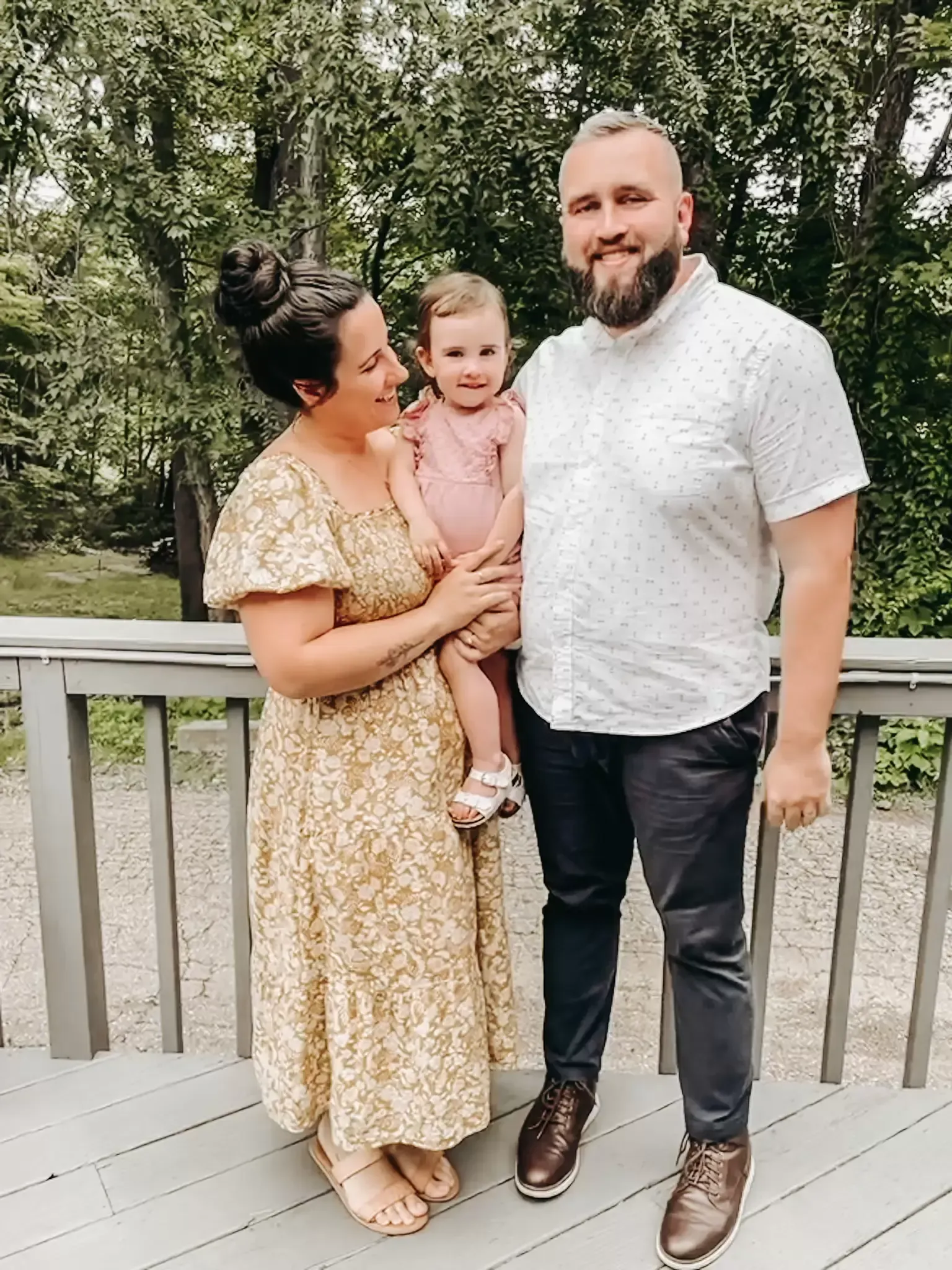 Family of three on a wooden deck. Mother holds a child; father stands. All smile.