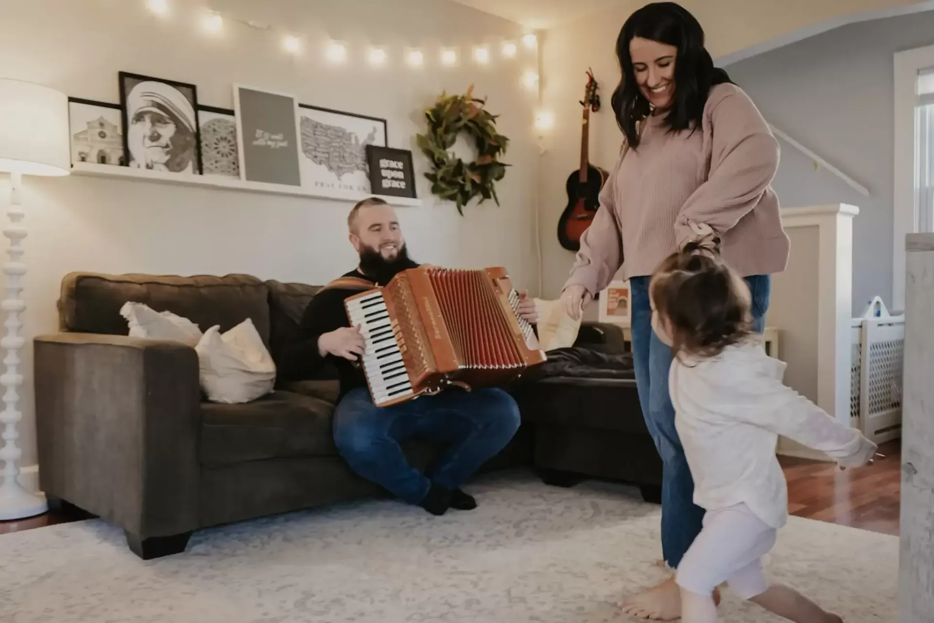 Man plays accordion, woman and toddler dance in cozy living room.