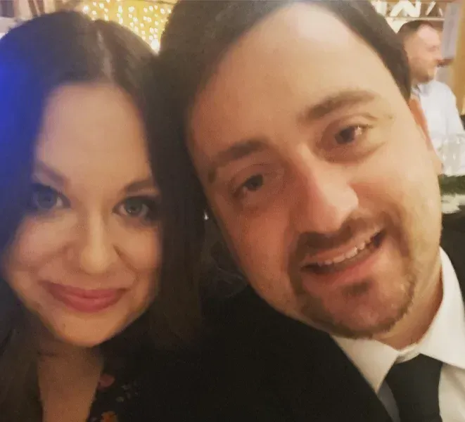 Woman and man smiling close together at an event. Dark clothing, happy expressions, warm lighting.