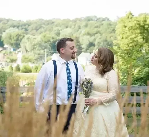 Bride and groom smile at each other outdoors. She holds bouquet, wears vintage dress. He wears suspenders and tie.