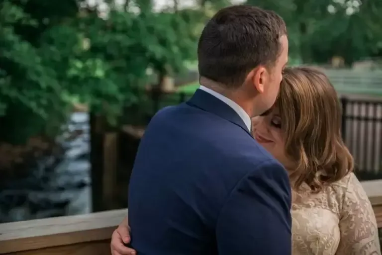 Groom in blue suit embraces bride in lace dress. They are outdoors on a wooden deck by a stream.