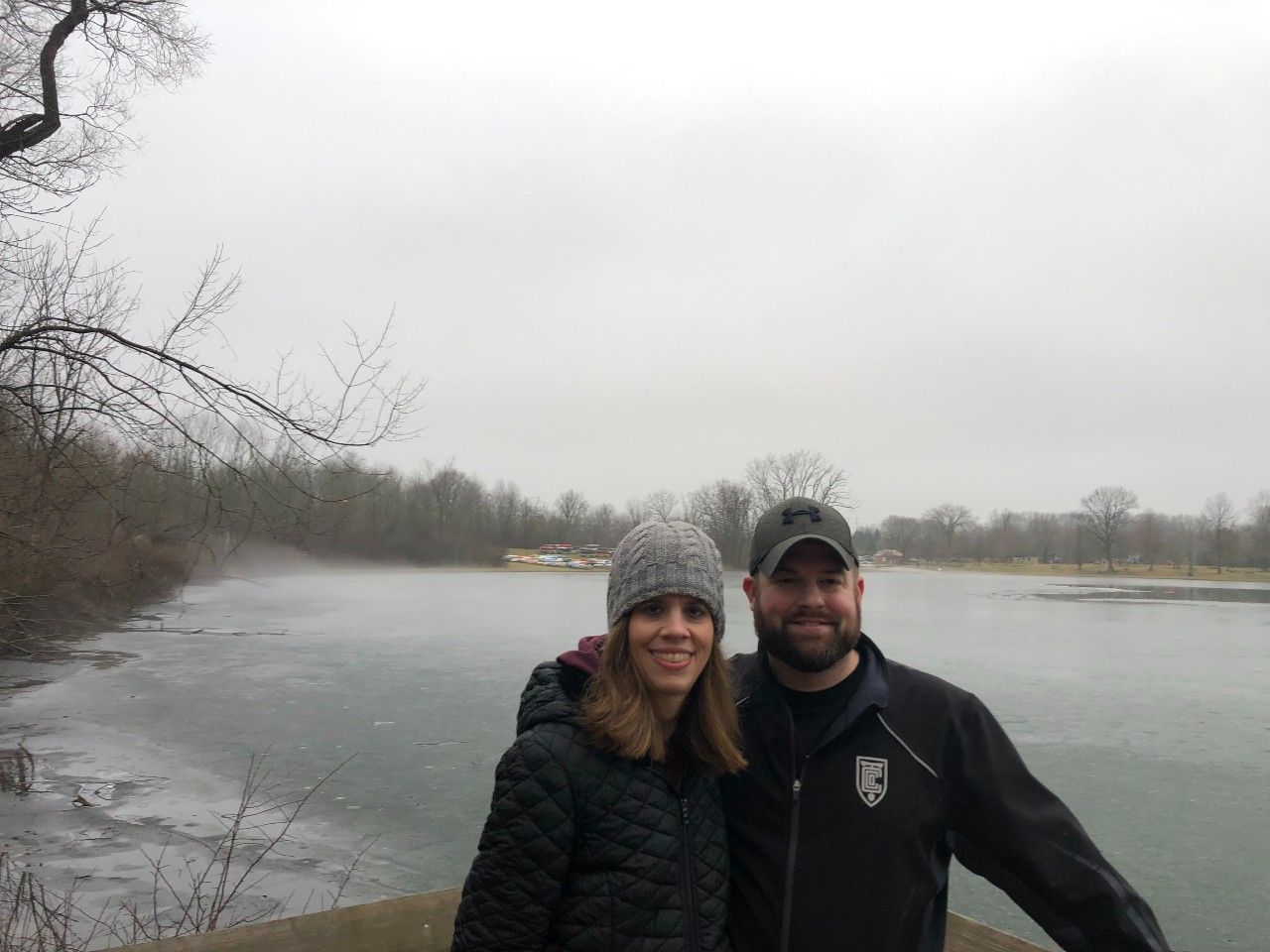 Couple smiles, posing by a misty lake. Woman in hat, man in cap, overcast sky.