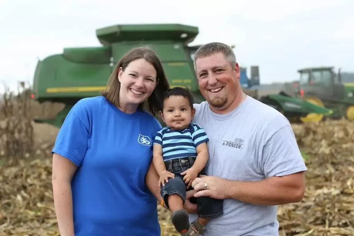 Family of three smiling, posing in a field with farm equipment.