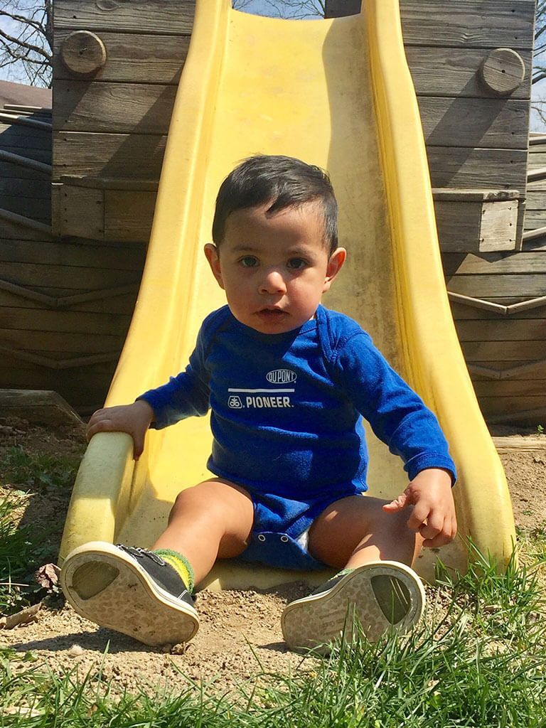 Young child sits on a yellow slide outdoors, wearing blue and black shoes, blue top and looking at the camera.