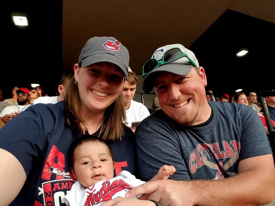 Family at a baseball game, smiling. Woman and man wearing hats and shirts with team logo, holding a baby.