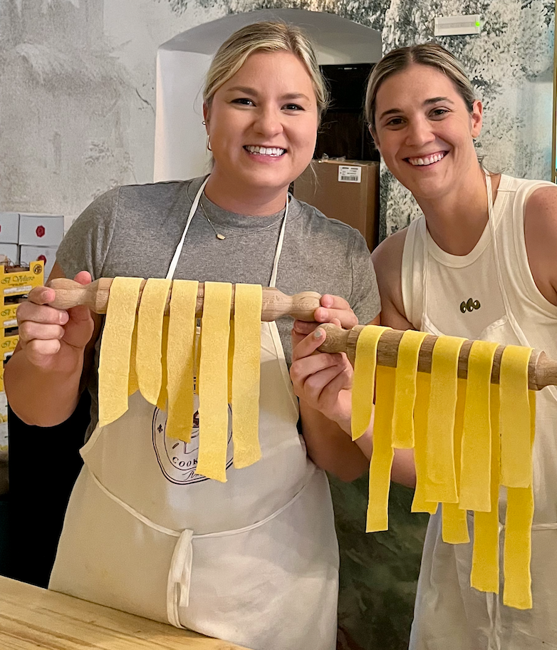Two women smiling, holding rolling pins with fresh pasta strips in a kitchen.