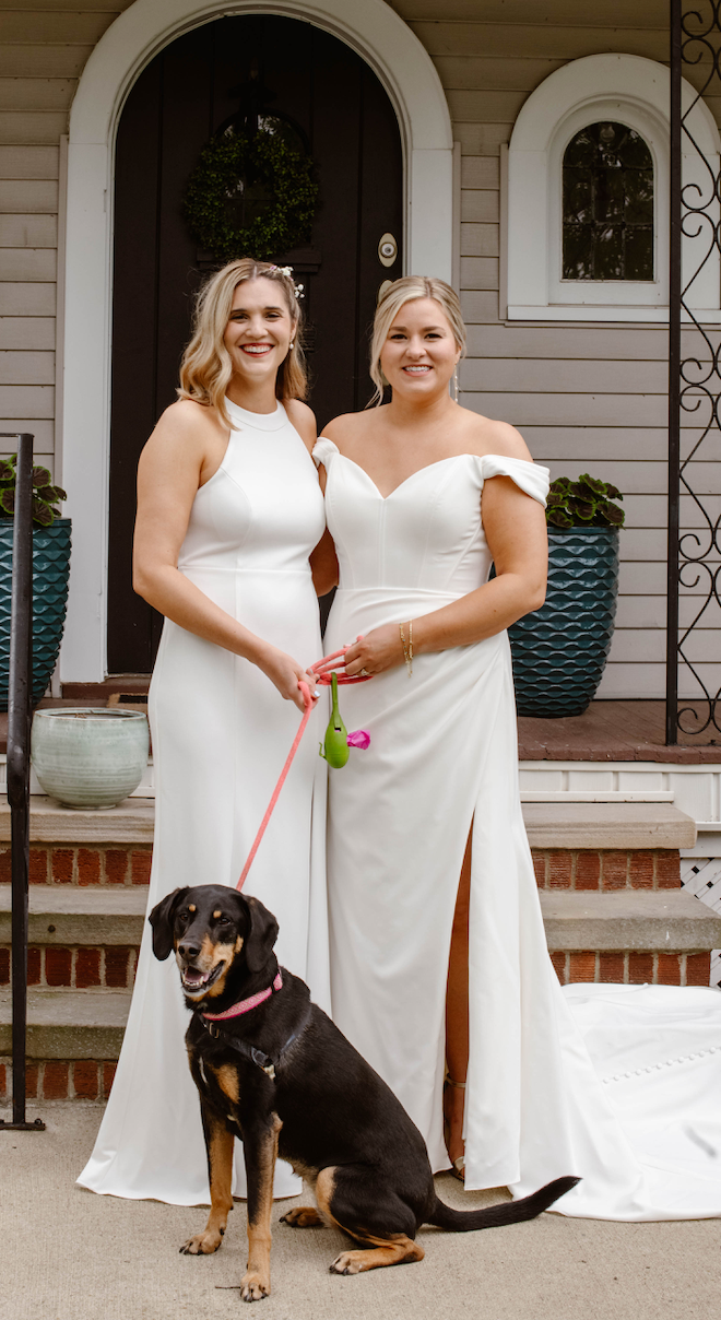 Two women in white wedding dresses with dog outside house.