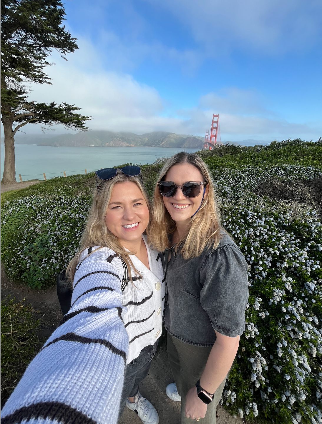 Two women smiling, taking a selfie near the Golden Gate Bridge, sunny day.