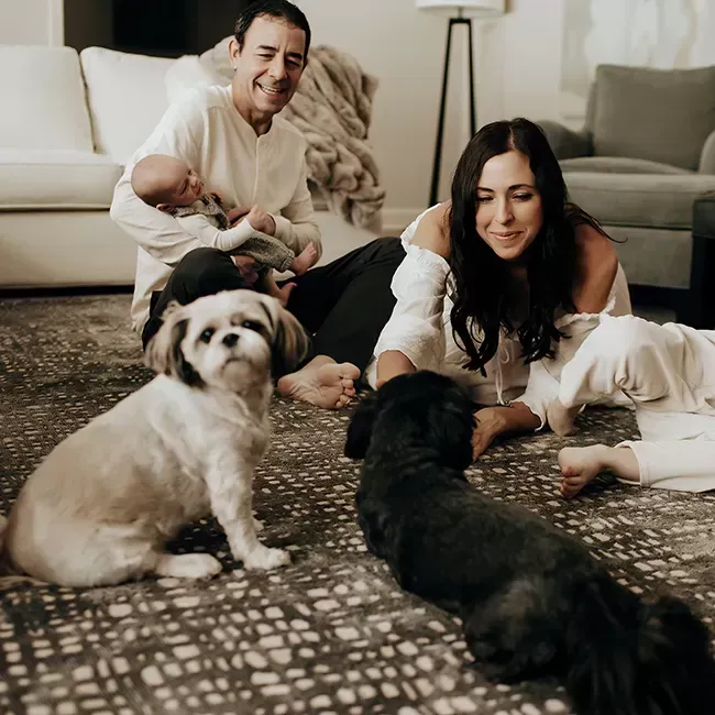 Family on floor, cuddling newborn and dogs. Man holds baby, woman smiles, two dogs look up.