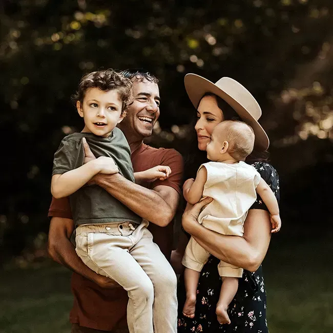 Family portrait: Father, mother, two children outdoors. Dad holds son, mom holds baby. All are smiling.