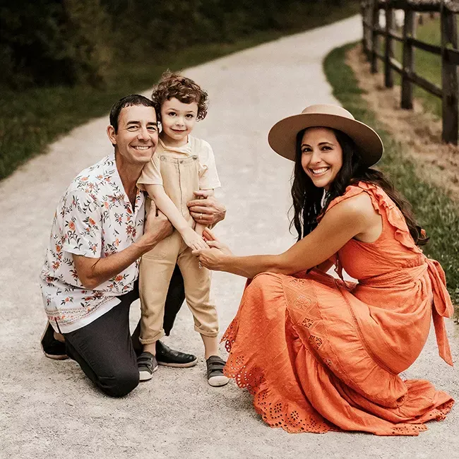 Family of three on a path. Man kneels, woman squats, both holding child's hands. All smiling, wearing summer attire.