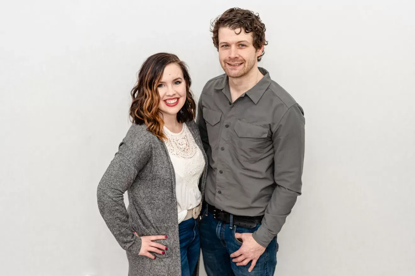 Woman with hand on hip, smiling, beside a man with a hand in pocket, smiling, in front of a white backdrop.