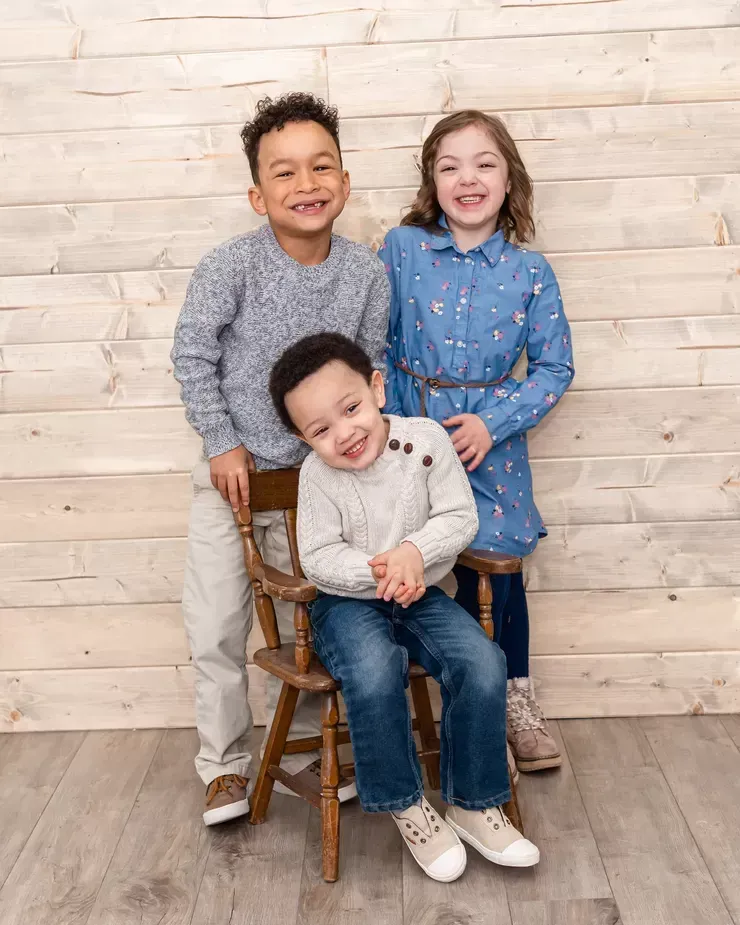 Three smiling children pose in front of a wooden backdrop. One sits on a chair, two stand behind.