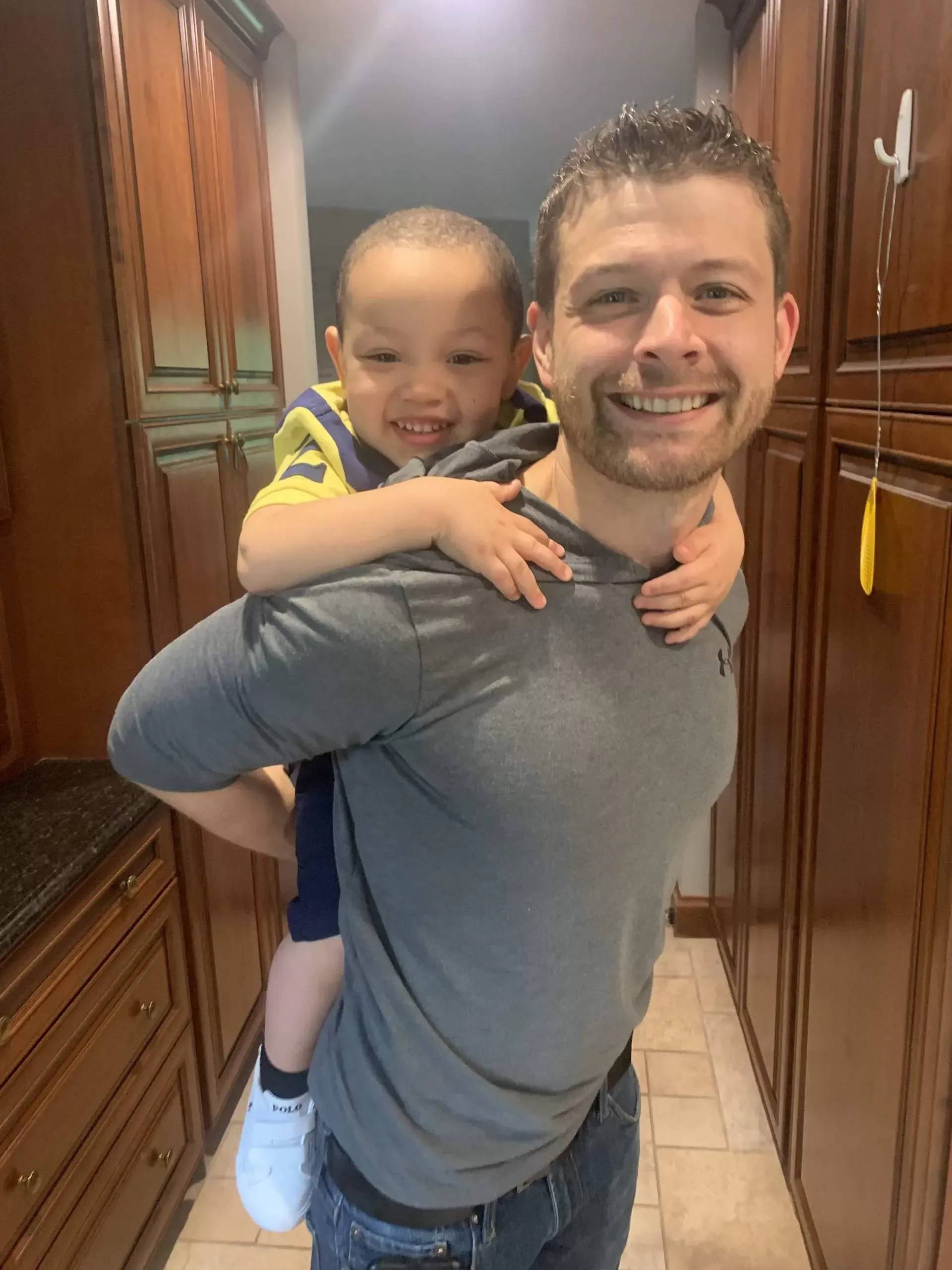 Man smiling, giving a piggyback ride to a smiling young child in a kitchen.