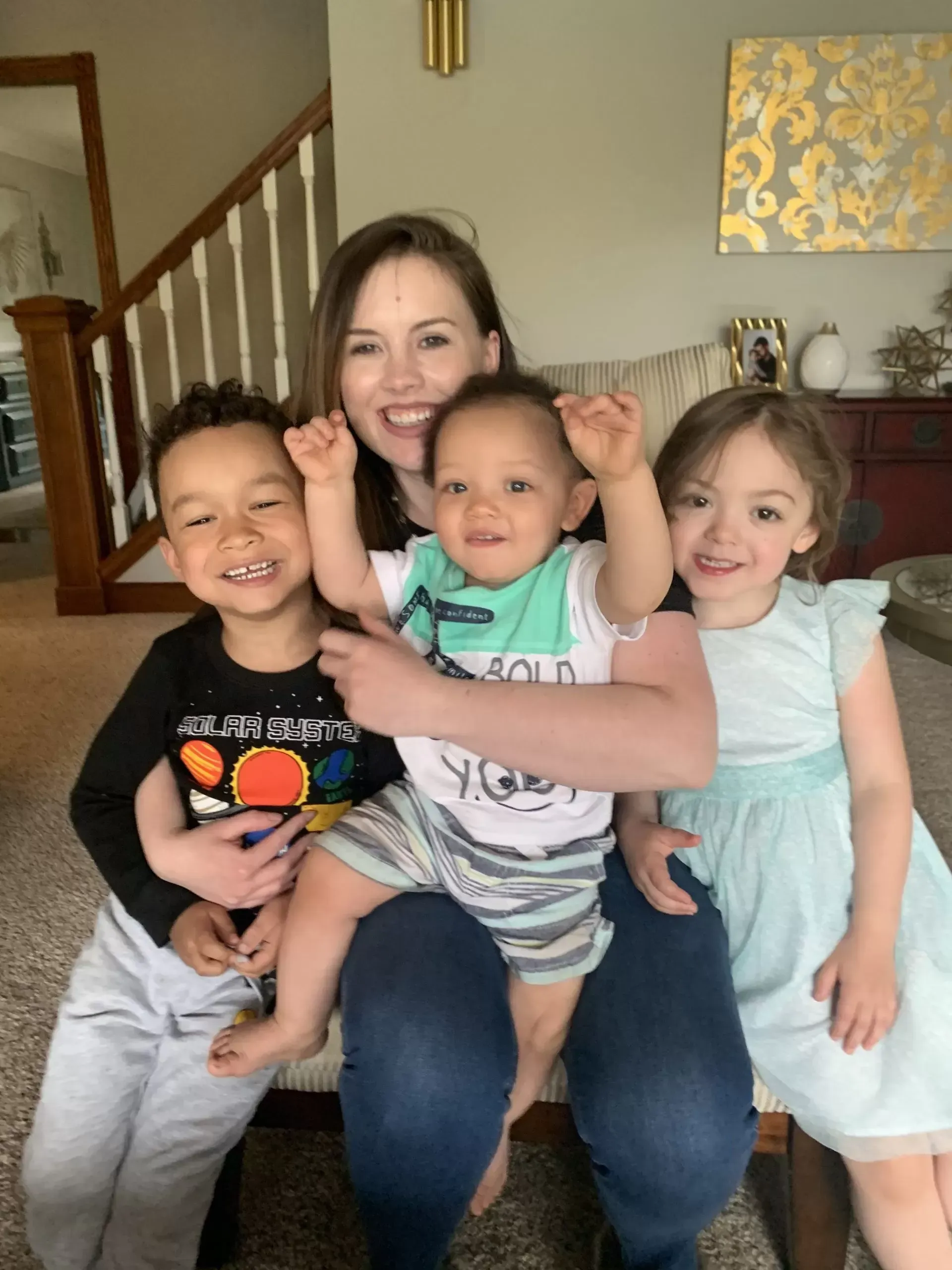 Woman smiles, holding three young children indoors; one raises arms, others smile.