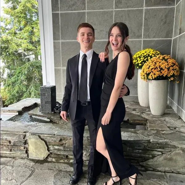 A couple, formal attire, smiles on a stone patio with potted yellow flowers.