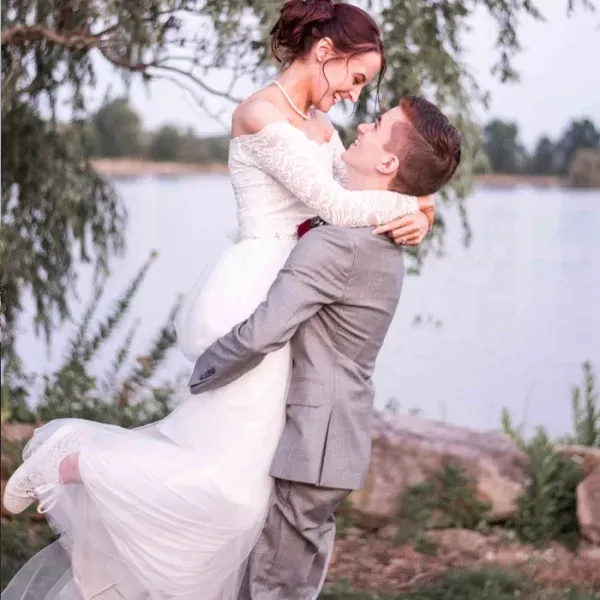 Bride in white dress, groom in gray suit, embrace near a lake. They are smiling, he's lifting her up.