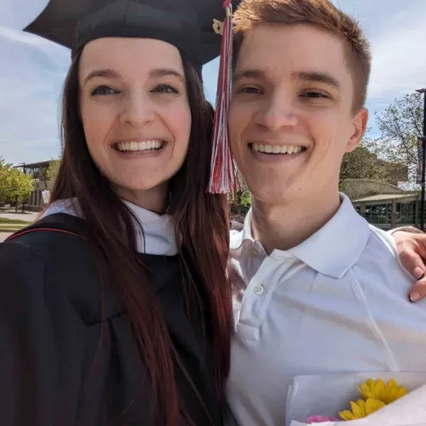 Woman in graduation cap and gown smiles with a man in a white polo shirt outdoors; both are smiling.