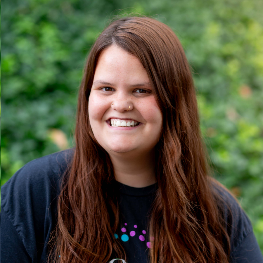 Woman with long brown hair smiling, in front of greenery. She wears a black shirt with a colorful design.
