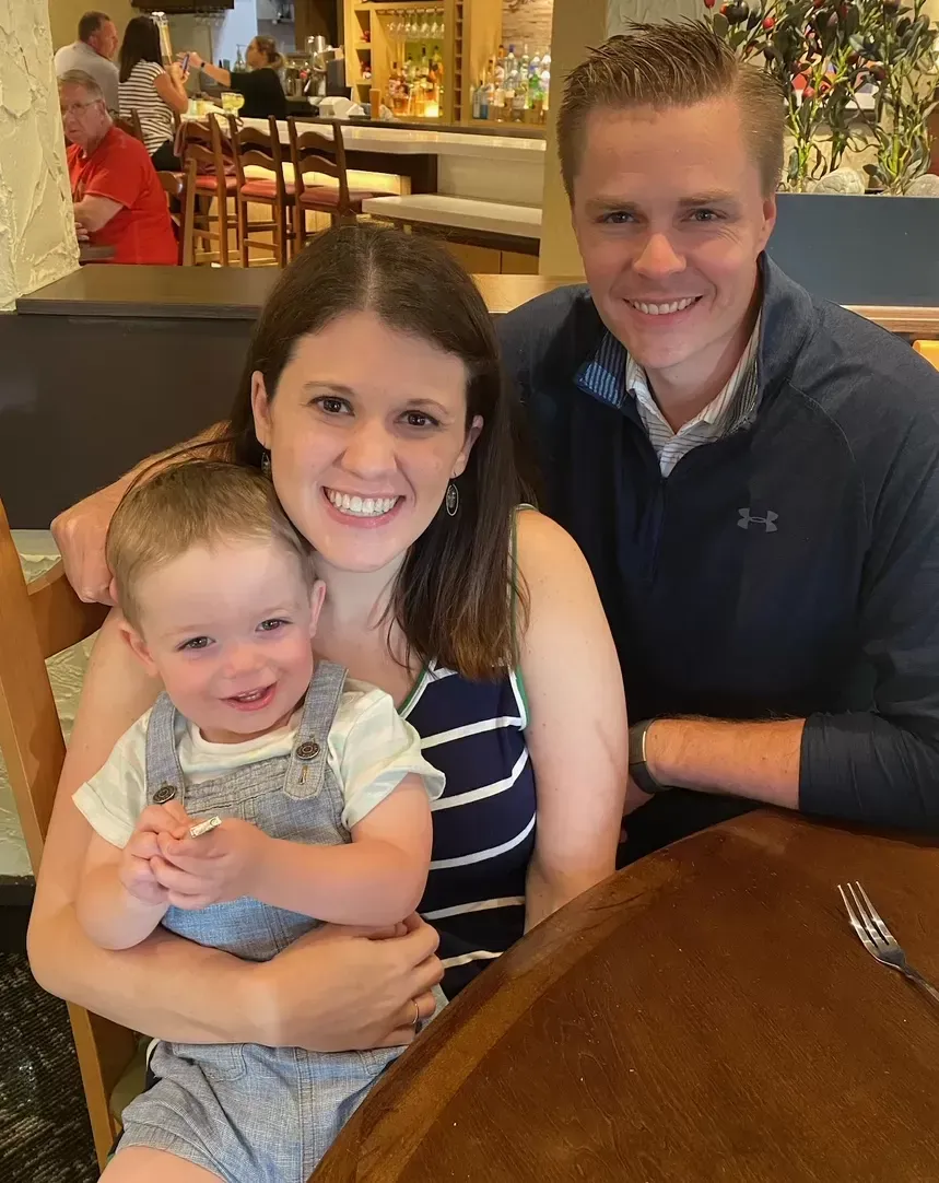 Family of three smiles at camera at a restaurant. Mom holds toddler, dad sits next to them.