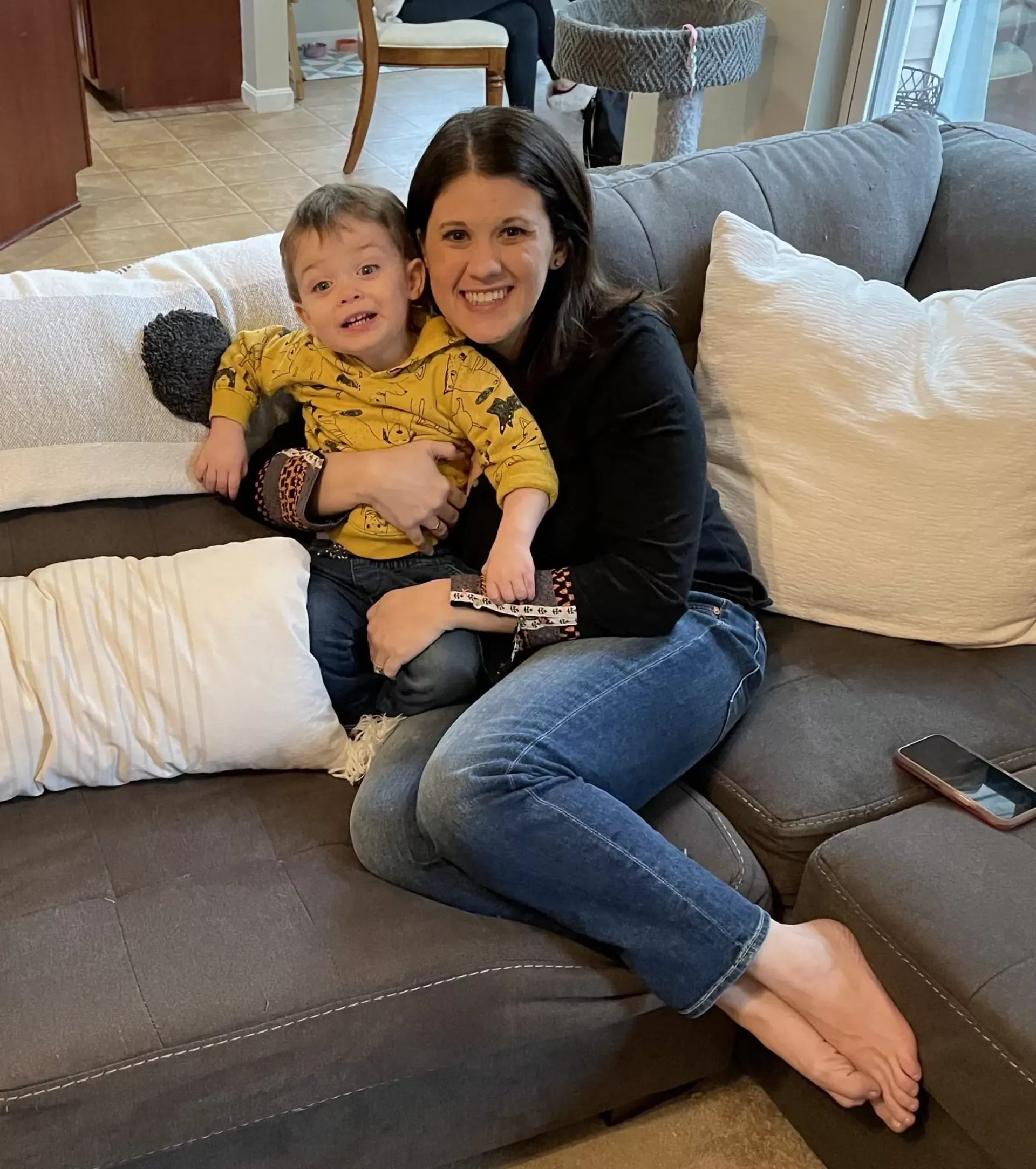 Woman and child sitting on a gray couch, smiling. Child wears a yellow hoodie.