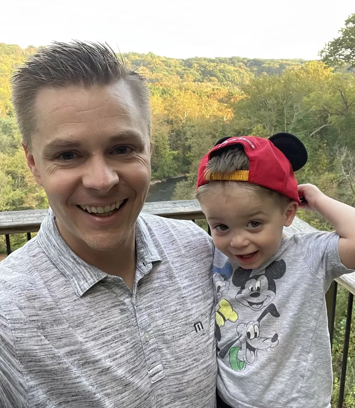 Man smiling, holding a young child wearing a Mickey Mouse hat and shirt, on a deck with a wooded view.