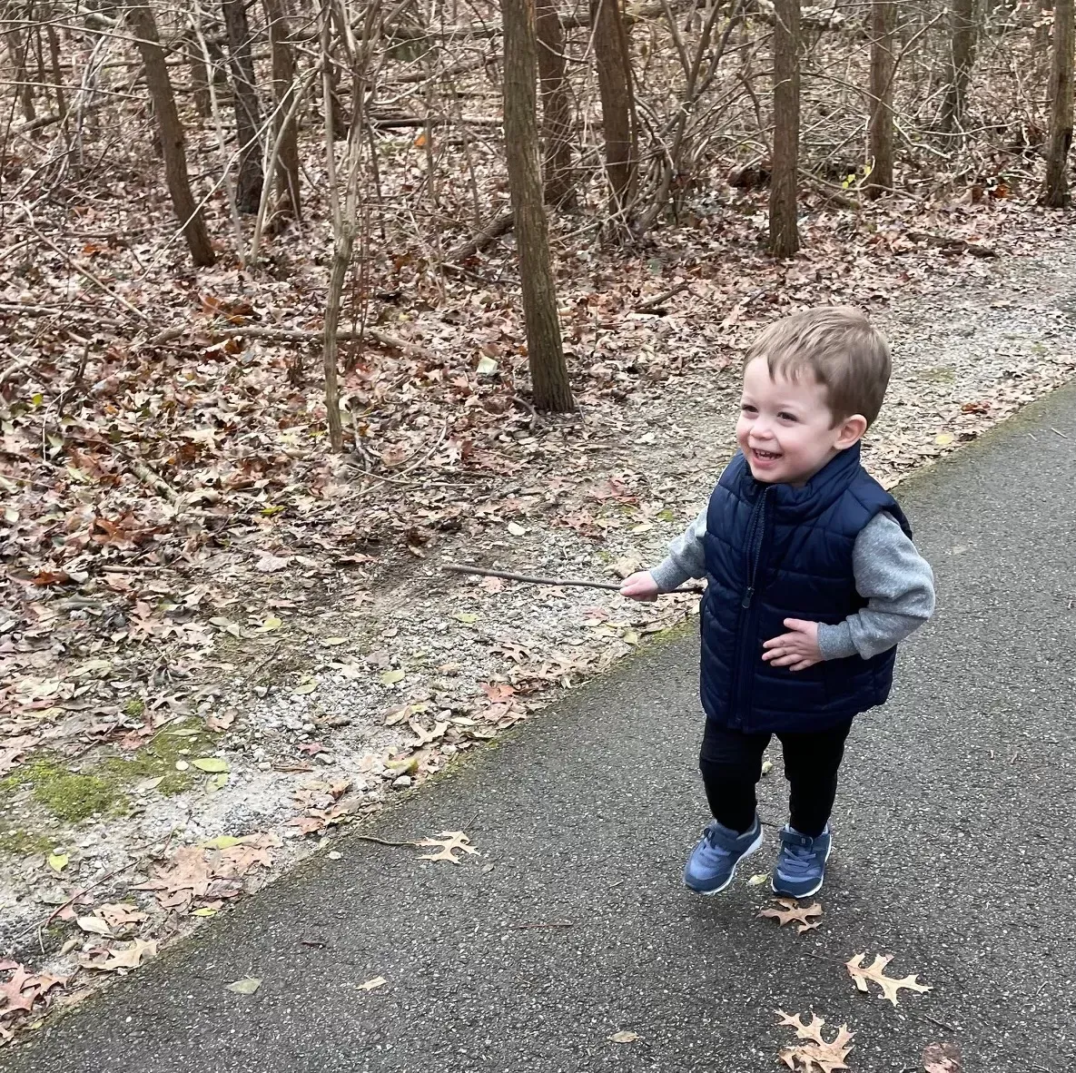 Smiling toddler in a blue vest runs on a paved path in a wooded area.