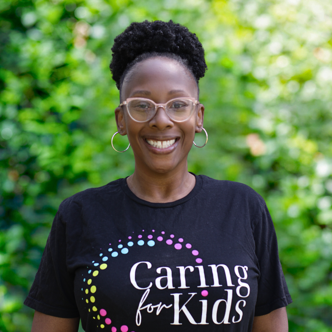 Woman with glasses smiles; wearing black sweatshirt, gray hair with curly braids, neutral background.