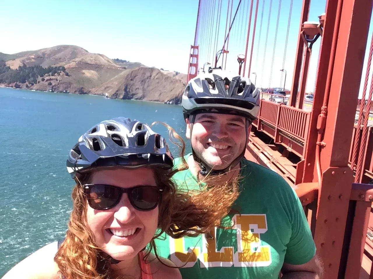 Couple on bikes, smiling on the Golden Gate Bridge, blue water, red bridge.