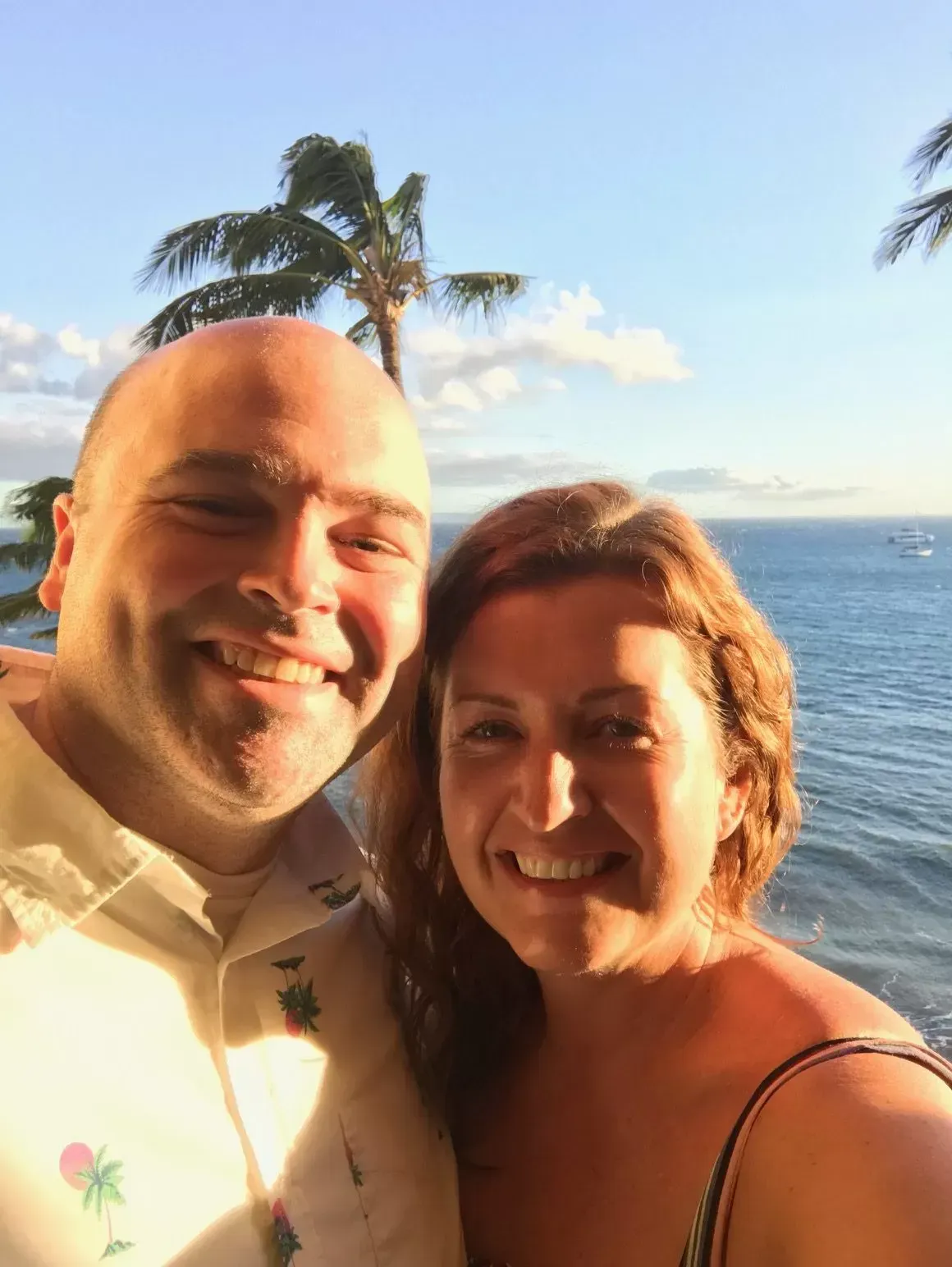 Couple smiling for selfie on a beach with ocean and palm trees in the background.