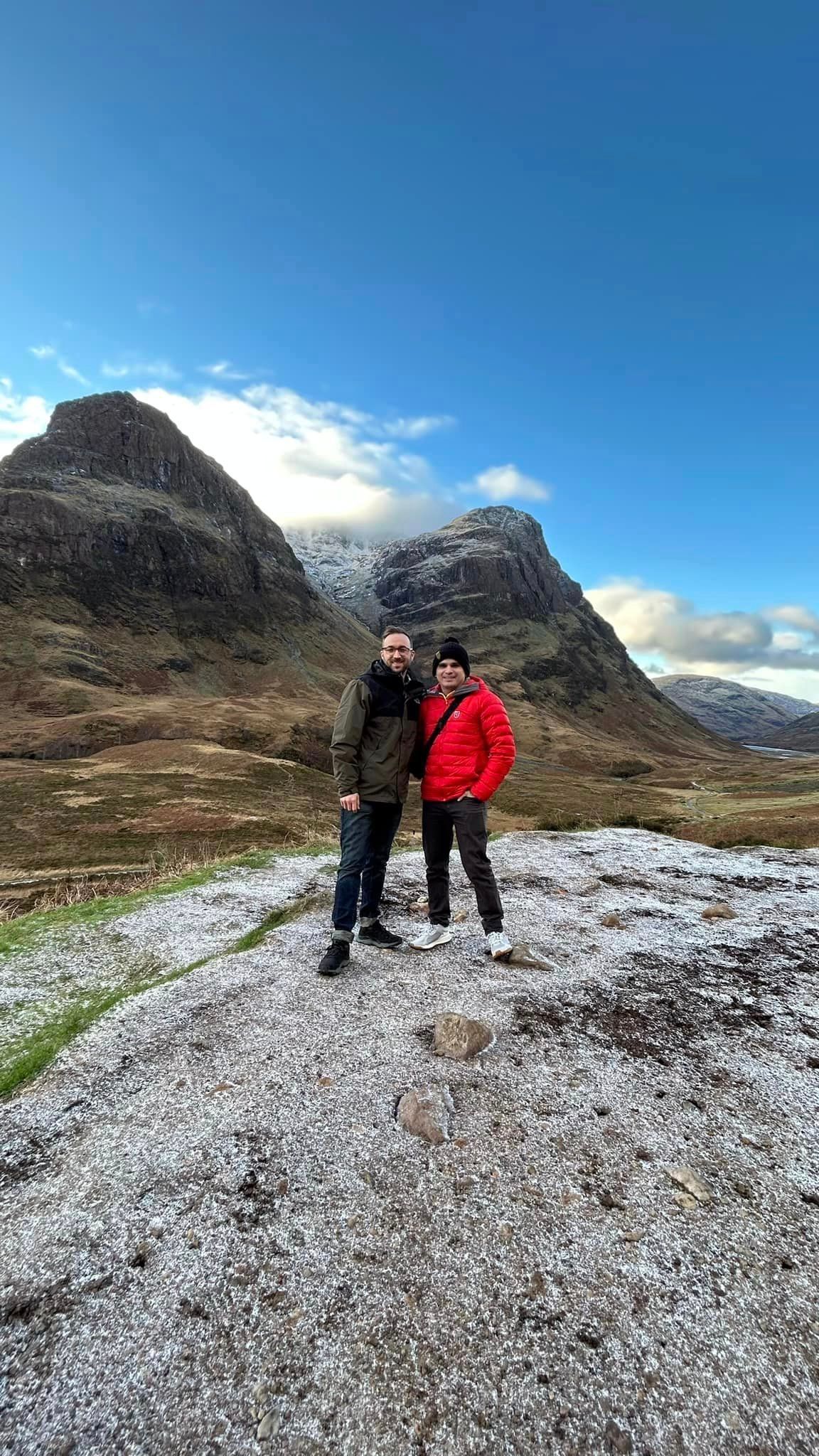Two people pose on a snowy hill, mountains behind them under a blue sky.