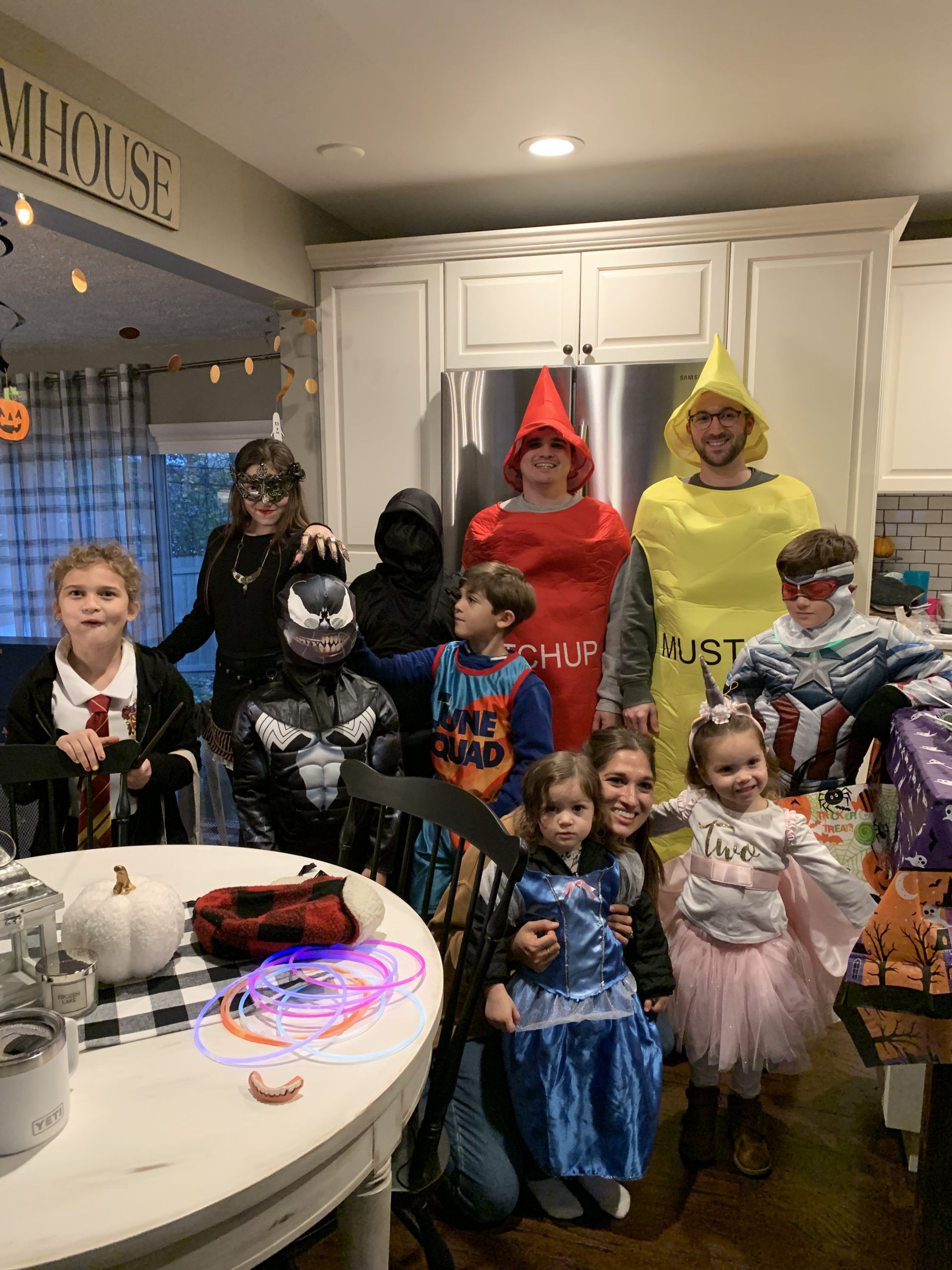 Group of people in Halloween costumes posing in a kitchen. Characters include Harry Potter, Venom, and crayons.