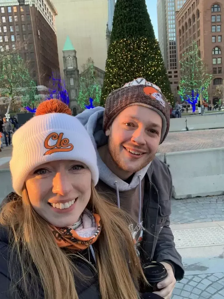 Two people wearing sports hats smile in front of a Christmas tree in a city setting.