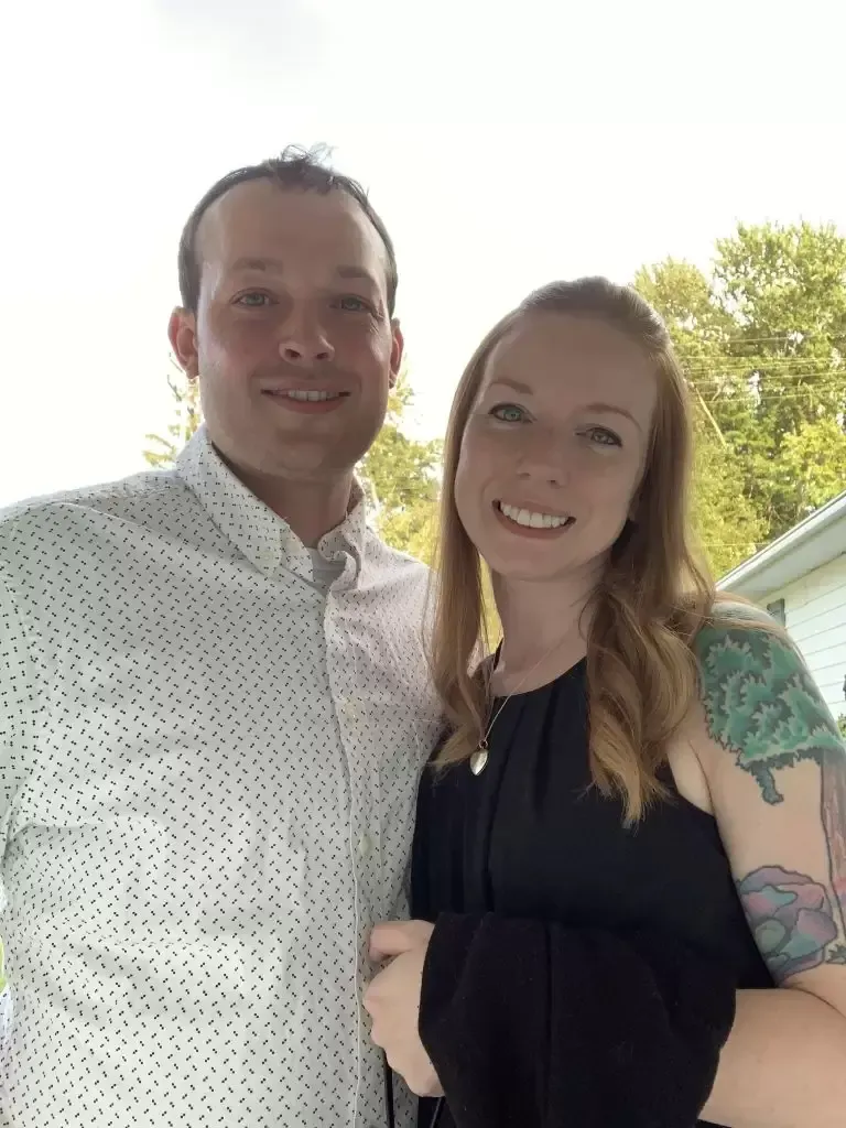 Man and woman smiling, posed together outdoors. The woman has a tattoo on her arm.