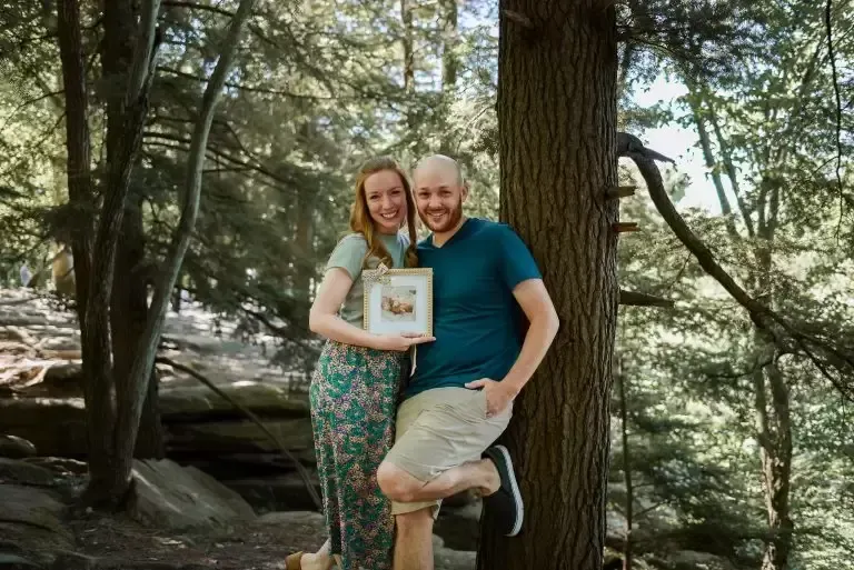 Couple in a forest, smiling, holding a framed photo, woman in floral pants and man in teal shirt and shorts.