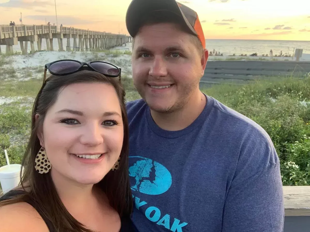 Couple smiling for a selfie on a beach at sunset, with a pier in the background.