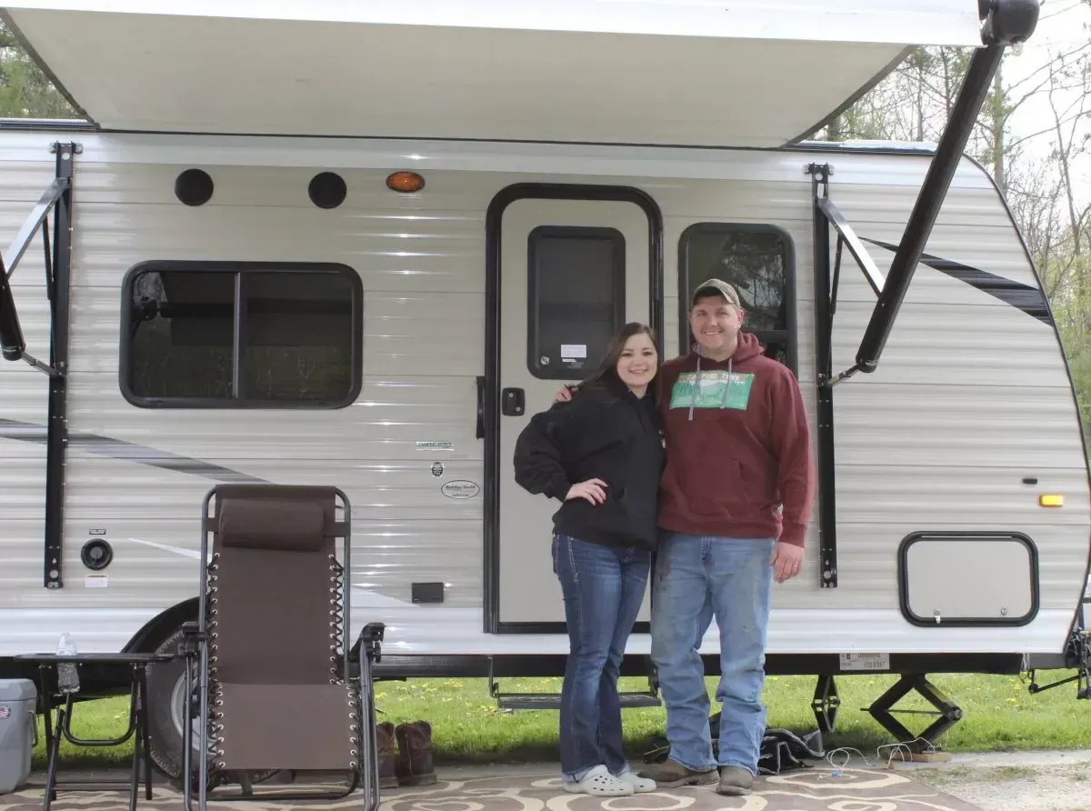 A couple stands in front of a camper. The man wears a maroon sweatshirt.