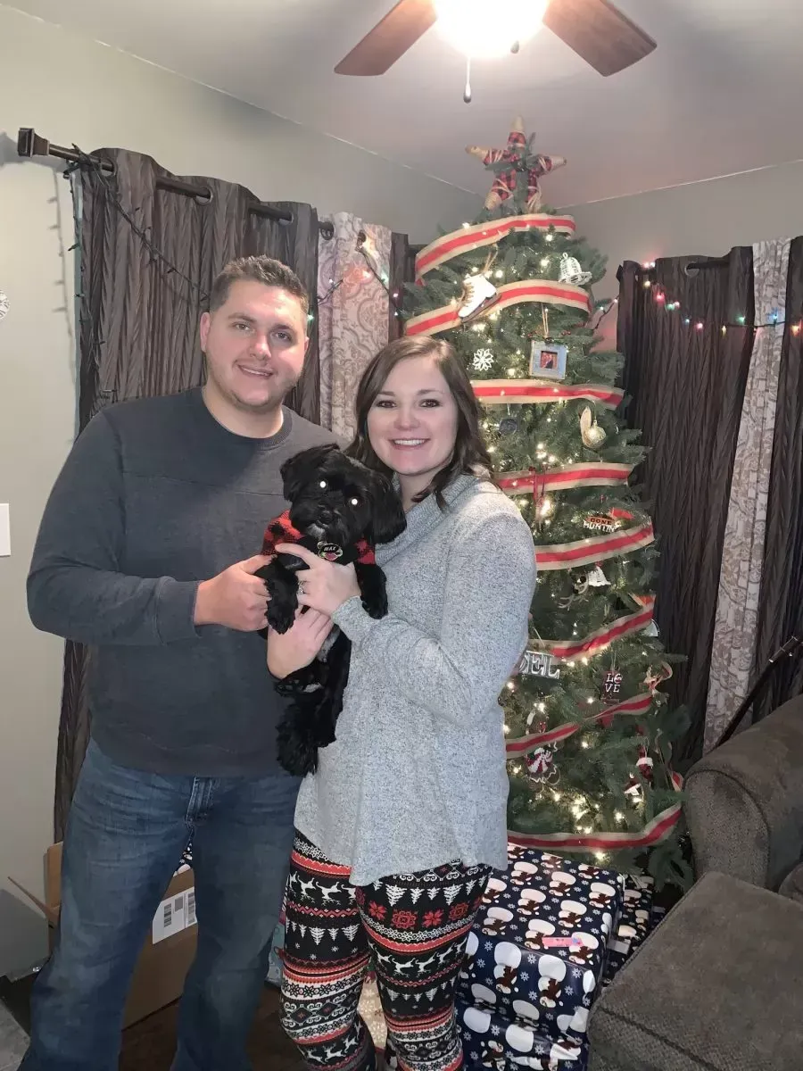Couple with a black dog in front of a Christmas tree; woman holding the dog is wearing holiday pajama pants.