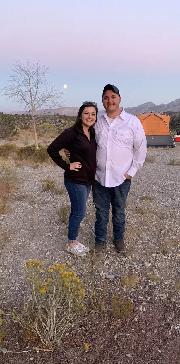 Couple posing outdoors with sunset sky; man in white shirt, woman in black; gravel ground.